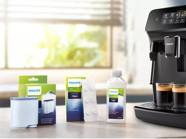 Na blacie kuchennym znajdują się akcesoria do ekspresu do kawy marki Philips, w tym filtr, tabletki czyszczące i płyn odkamieniający. Obok stoi ekspres do kawy z dwoma filiżankami espresso.