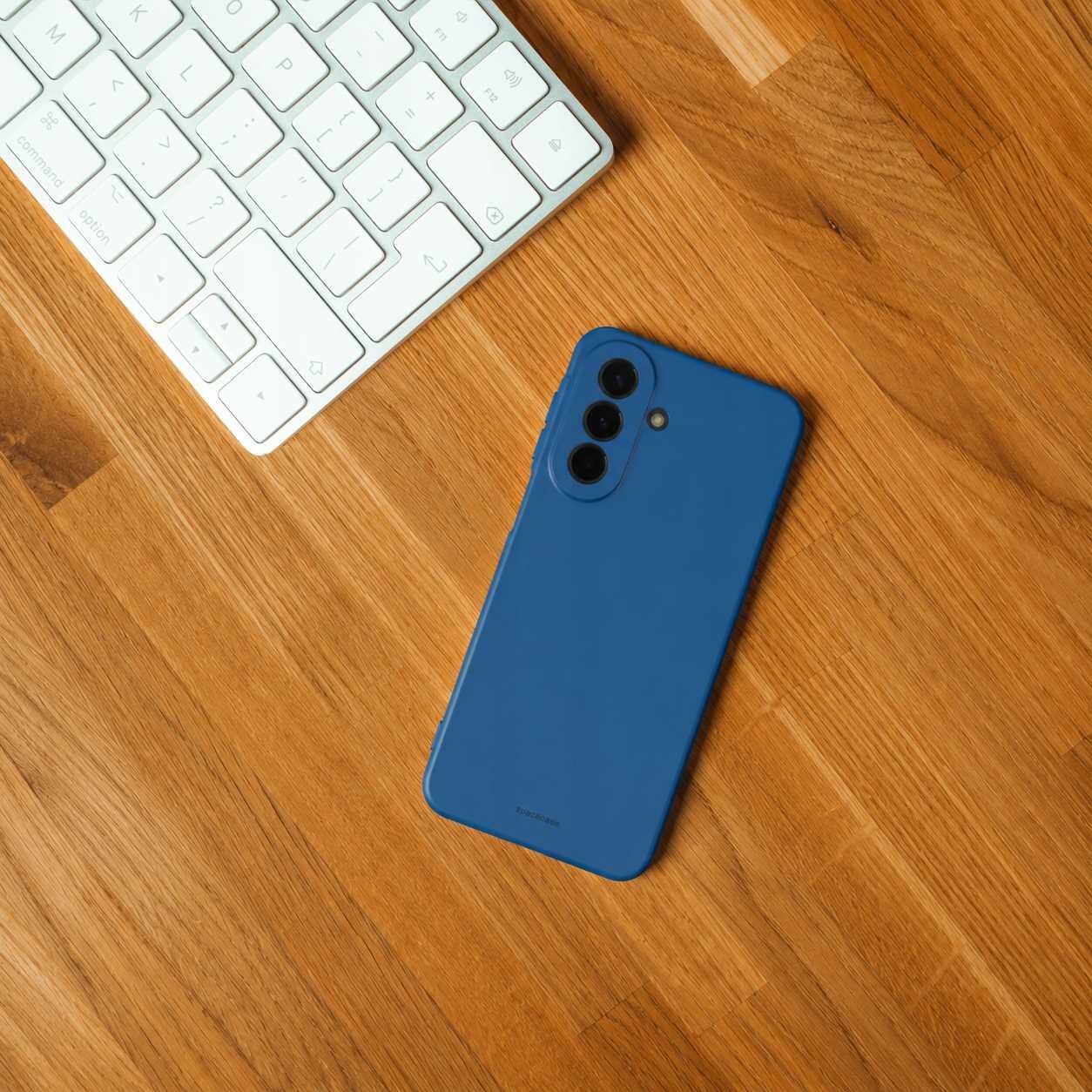 Na drewnianym blacie leży niebieskie silikonowe etui na telefon z trzema otworami na aparaty. Obok znajduje się biała klawiatura komputerowa.