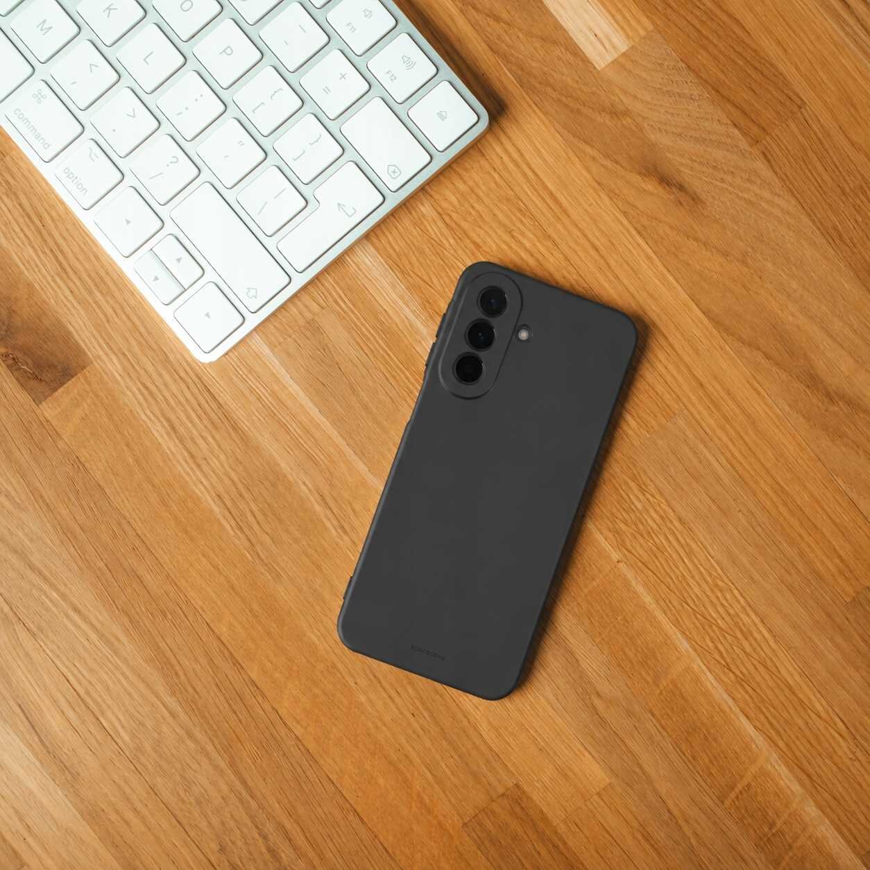 Na drewnianym blacie leży czarny telefon w silikonowym etui obok białej klawiatury. Etui ma wycięcia na trzy obiektywy aparatu i lampę błyskową.
