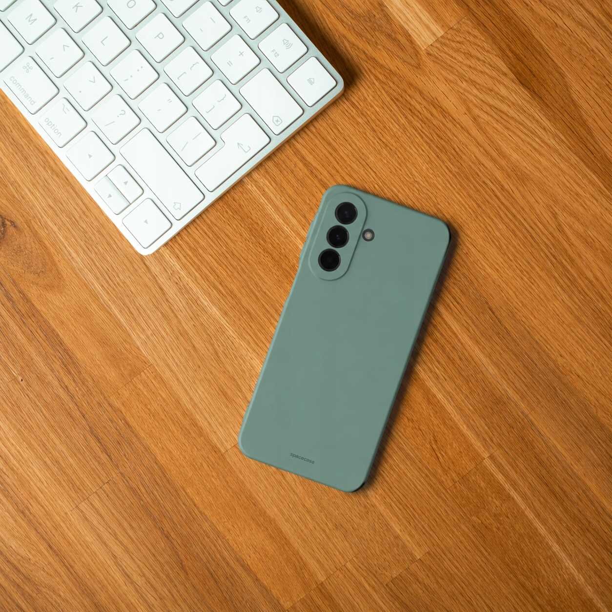Na drewnianym blacie leży zielony telefon w silikonowym etui obok białej klawiatury. Etui ma wycięcia na trzy obiektywy aparatu i logo producenta na dole.