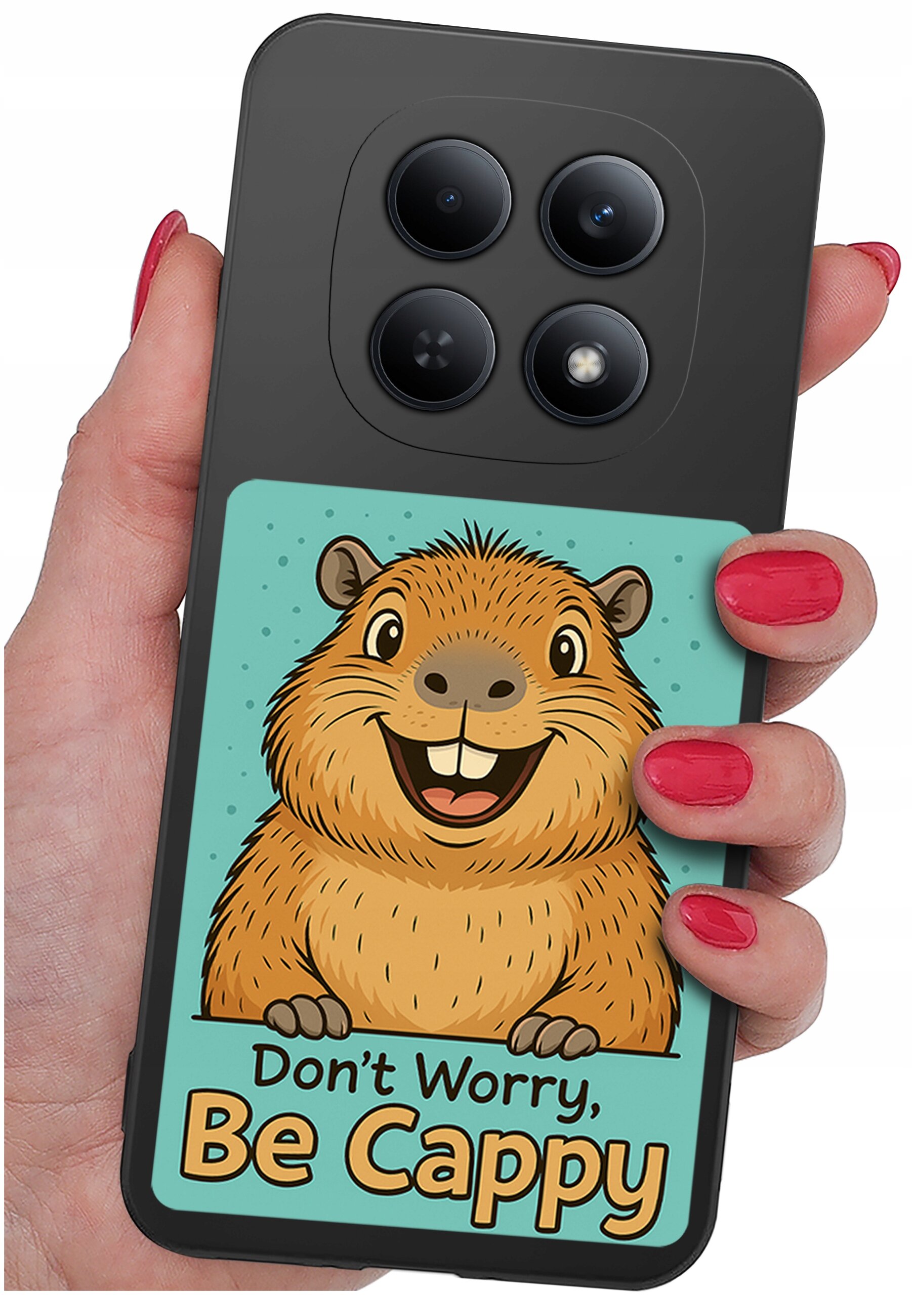 Osoba trzyma telefon z etui przedstawiającym rysunek kapibary z napisem \'Don\'t Worry, Be Cappy\'. Etui jest silikonowe i matowe, z czterema widocznymi obiektywami aparatu.