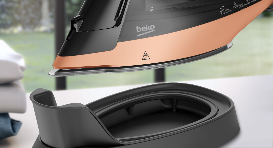 Na obrazie widoczne jest żelazko z logo \'beko\' w kolorze czarno-miedzianym, unoszące się nad podstawą. W tle widać rozmyty widok na zewnątrz przez okno.