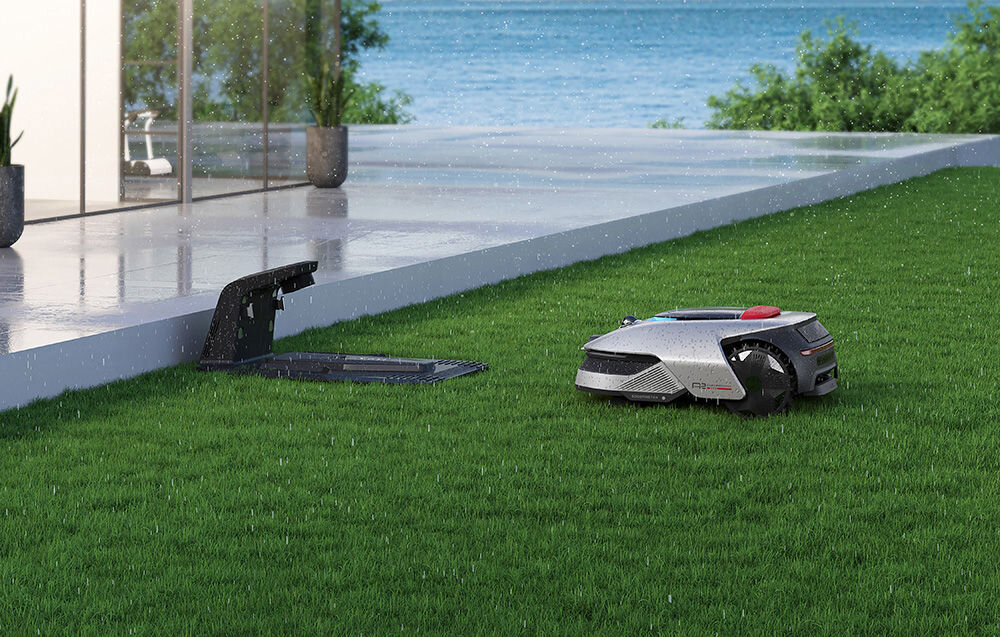 Na trawniku znajduje się robot koszący obok otwartej stacji dokującej. W tle widać taras i wodę, a na robocie widoczny jest napis \'DREAME\'.