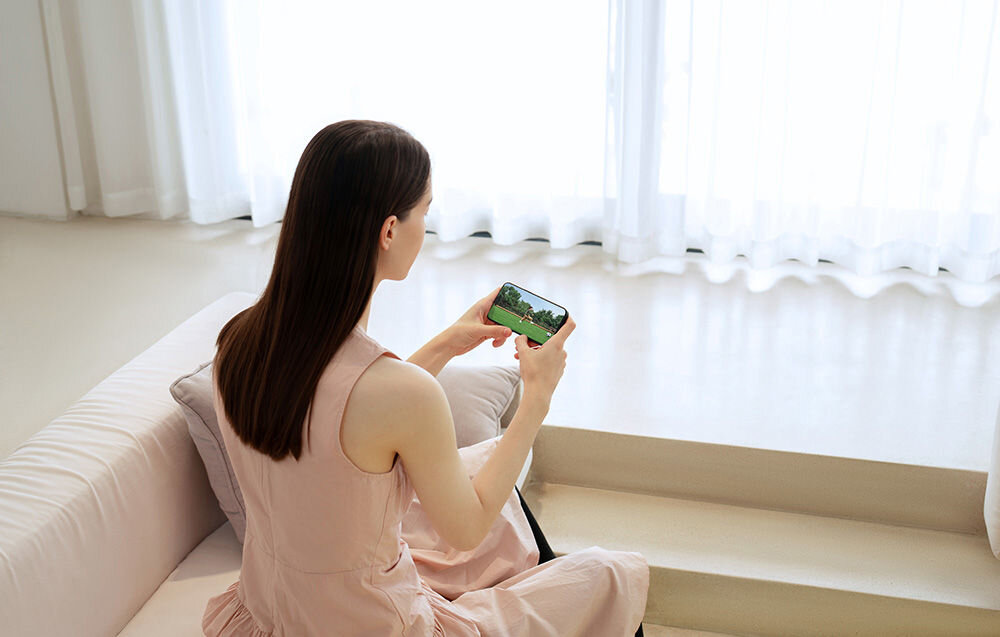 Osoba siedzi na kanapie i trzyma w rękach smartfon, na którym wyświetlany jest obraz zielonego krajobrazu. W tle widoczne są jasne zasłony przepuszczające światło.