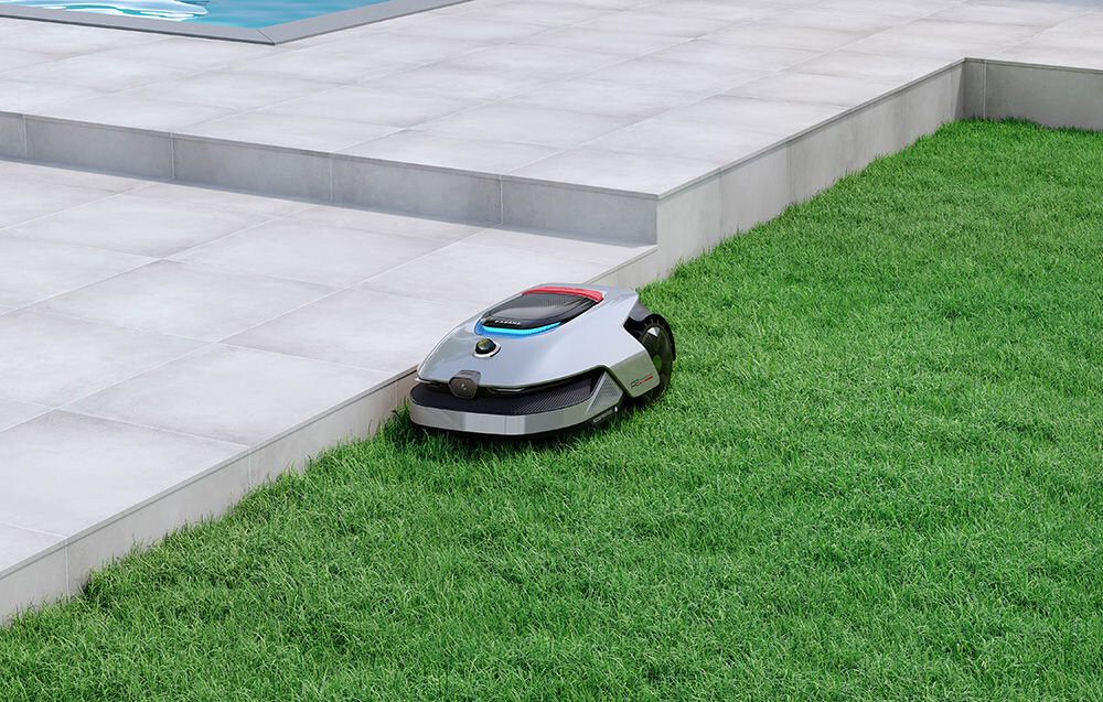 Robot koszący porusza się po trawniku obok wyłożonego płytkami tarasu. Urządzenie ma srebrną obudowę z czarnymi elementami i widocznym logo producenta.