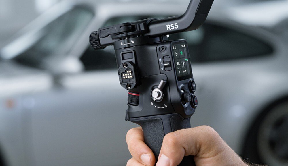 Osoba trzyma gimbal z widocznym napisem \'RS5\' oraz ekranem z ikonami. W tle rozmyty samochód.