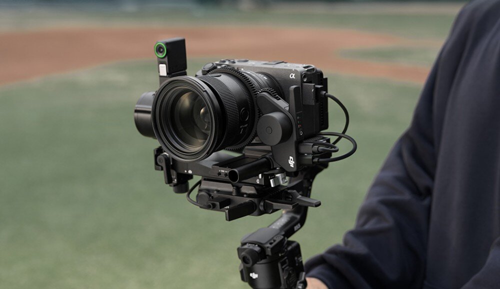 Osoba trzyma gimbal z zamontowanym aparatem fotograficznym. W tle widoczne jest rozmyte boisko sportowe.