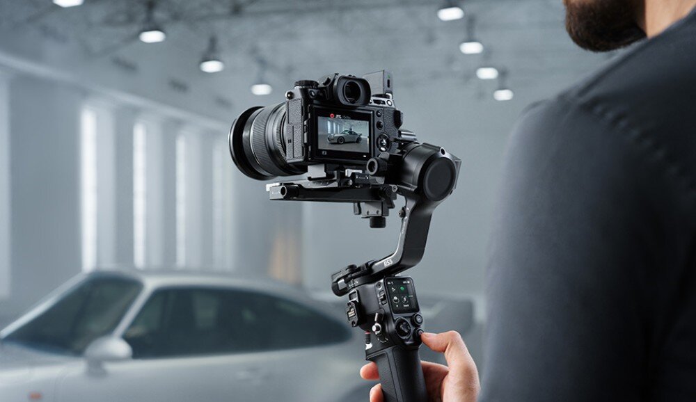 Osoba trzyma gimbal z zamontowanym aparatem, w tle widoczny jest samochód. Gimbal posiada wyświetlacz i kilka przycisków sterujących.