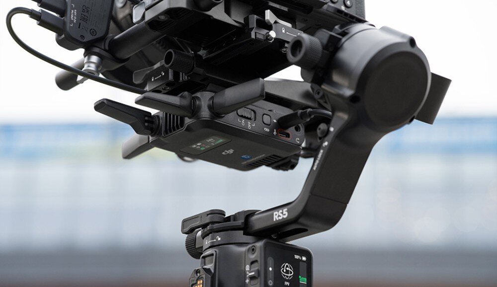 Na zdjęciu widoczny jest gimbal z oznaczeniem RS5, zamontowany na statywie. W tle znajduje się rozmyty budynek.