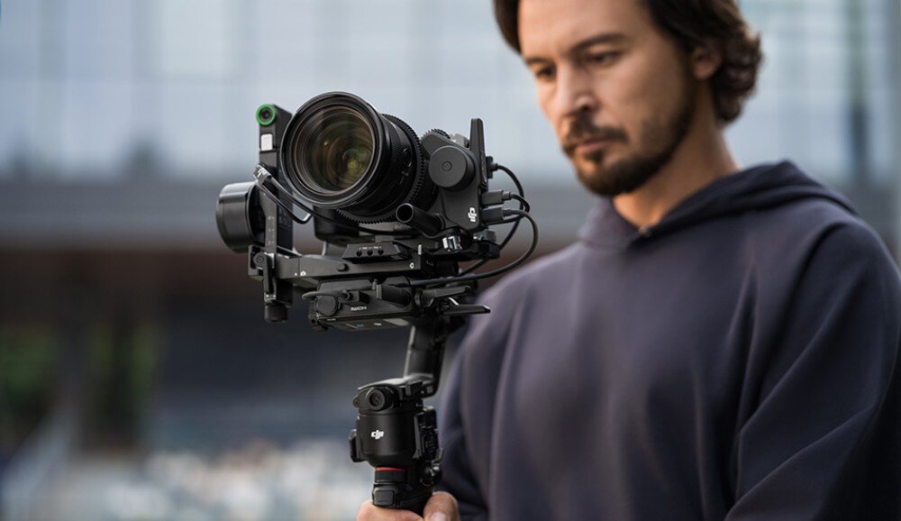 Osoba trzyma gimbal z zamontowanym aparatem fotograficznym. Na gimbalu widoczne jest logo DJI.
