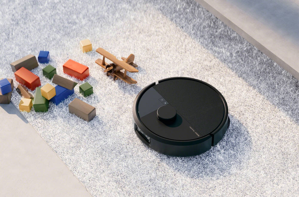 Na dywanie znajduje się czarny robot sprzątający oraz kolorowe klocki i drewniany model samolotu. Robot ma okrągły kształt i widoczne są na nim przyciski oraz logo producenta.