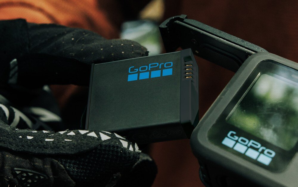 Osoba w rękawiczkach trzyma akumulator z widocznym logo GoPro obok kamery sportowej z wyświetlaczem.