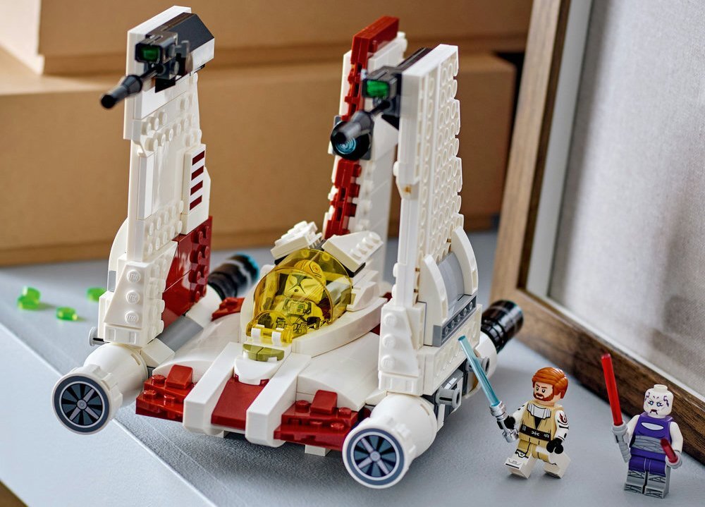 Na obrazie znajduje się model myśliwca z klocków LEGO z charakterystycznym żółtym kokpitem i czerwono-białymi elementami. Obok myśliwca stoją dwie figurki LEGO z mieczami świetlnymi.