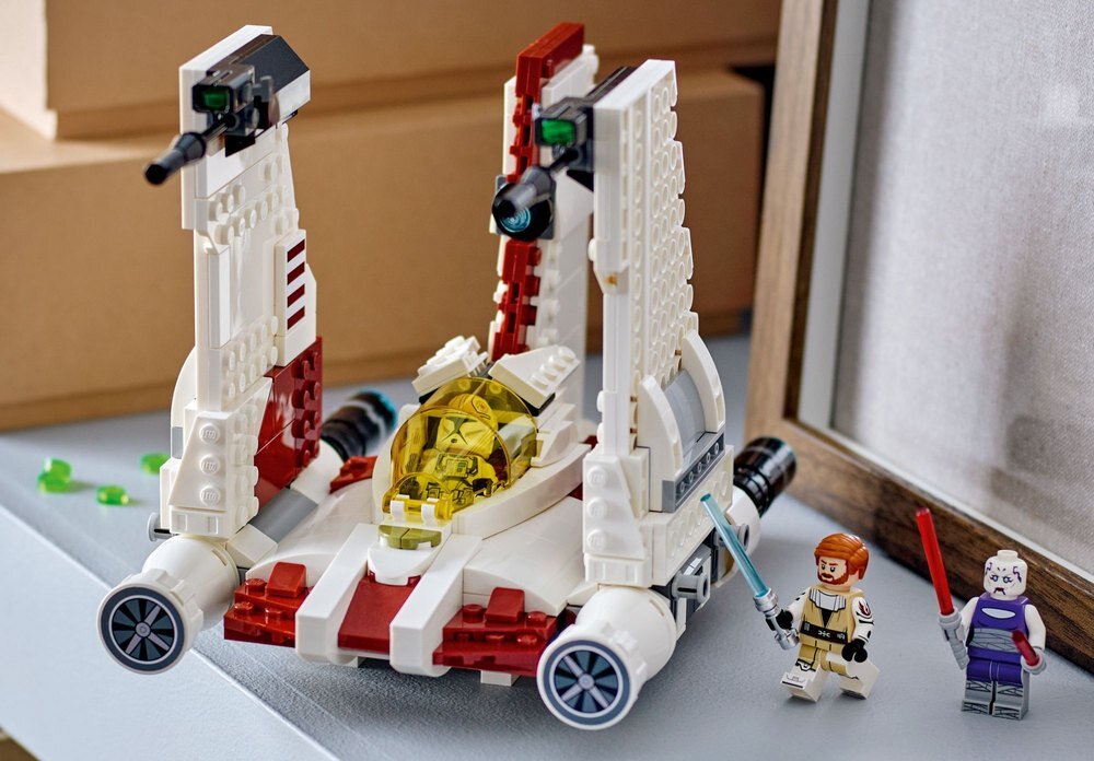 Na obrazie znajduje się model LEGO przedstawiający myśliwiec z dwoma figurkami postaci trzymającymi miecze świetlne. Myśliwiec ma białe i czerwone elementy oraz żółtą osłonę kokpitu.