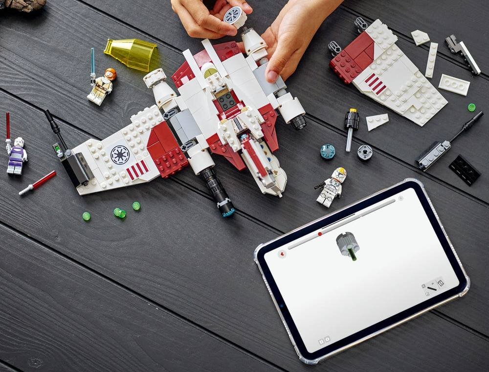 Na ciemnym drewnianym stole znajduje się zestaw klocków LEGO przedstawiający myśliwiec, z widocznymi figurkami postaci i elementami konstrukcyjnymi. Obok leży tablet z wyświetlonym schematem budowy klocków.