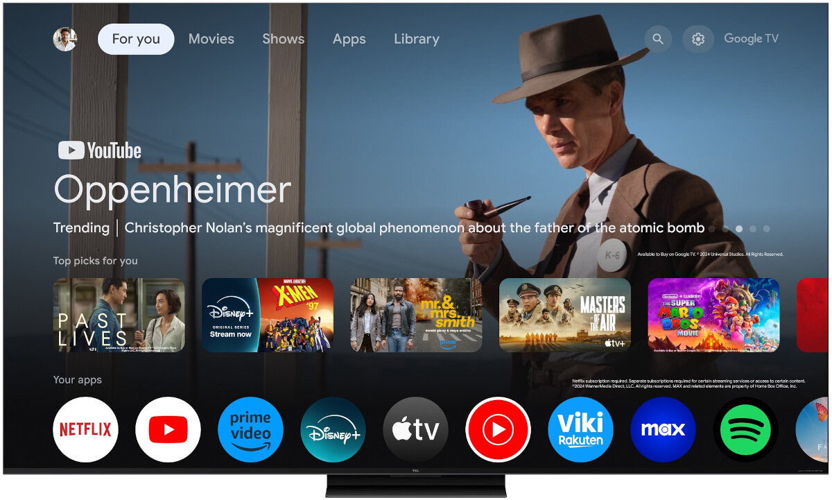 Ekran telewizora wyświetla interfejs Google TV z filmem \'Oppenheimer\' w tle. Na dole widoczne są ikony aplikacji, takie jak Netflix, YouTube, Prime Video, Disney+, Apple TV, Viki, Max i Spotify.