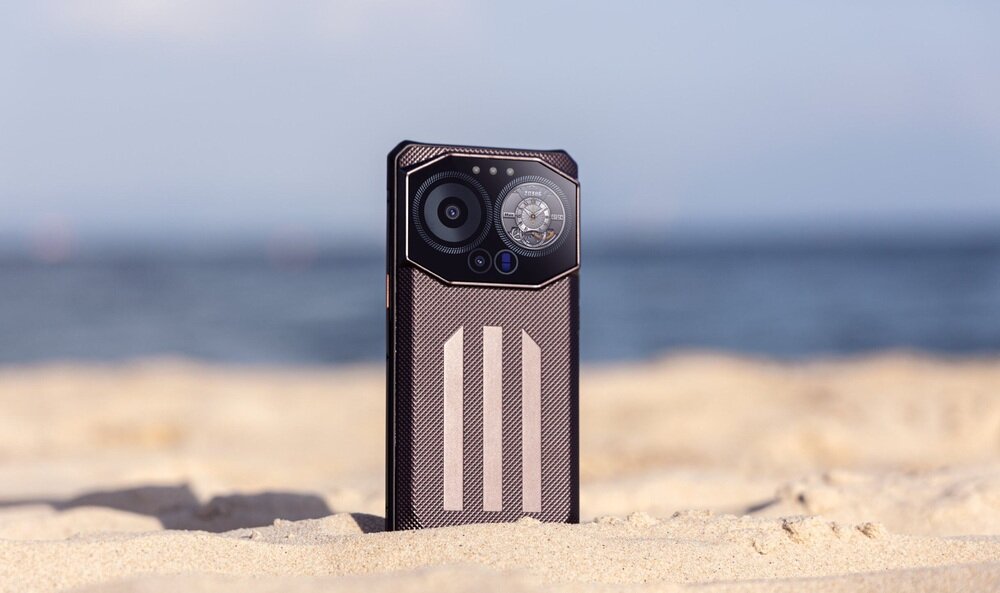 Smartfon stoi pionowo na piaszczystej plaży, w tle widać rozmyte morze i niebo. Obudowa telefonu ma wzór z trzema pionowymi paskami i widoczne są dwa obiektywy aparatu oraz okrągły element przypominający zegar.