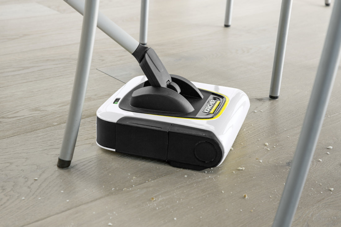 Elektryczna szczotka w kolorze białym z czarnymi elementami, z widocznym logo \'KARCHER\', znajduje się na podłodze wśród nóg krzesła. Na podłodze widoczne są drobne zanieczyszczenia.