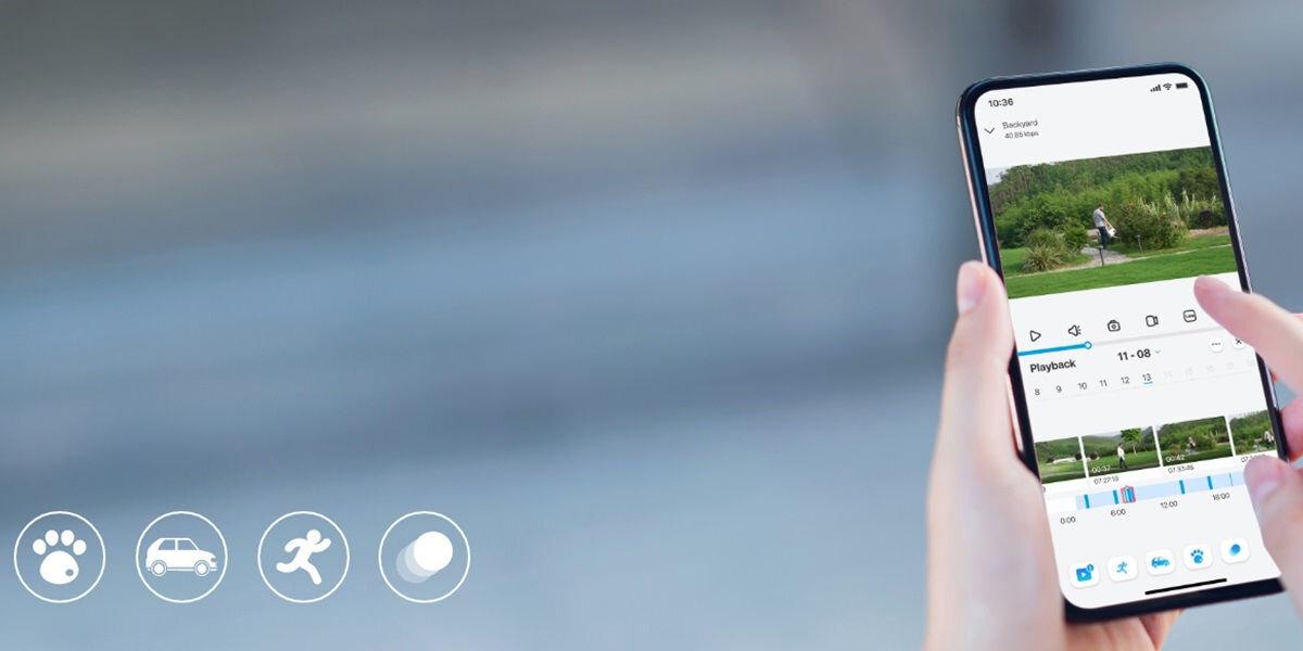Osoba trzyma smartfon, na którym wyświetlana jest aplikacja z podglądem z kamery i opcjami odtwarzania. W lewym dolnym rogu widoczne są ikony: łapa, samochód, biegnąca postać i jajko.