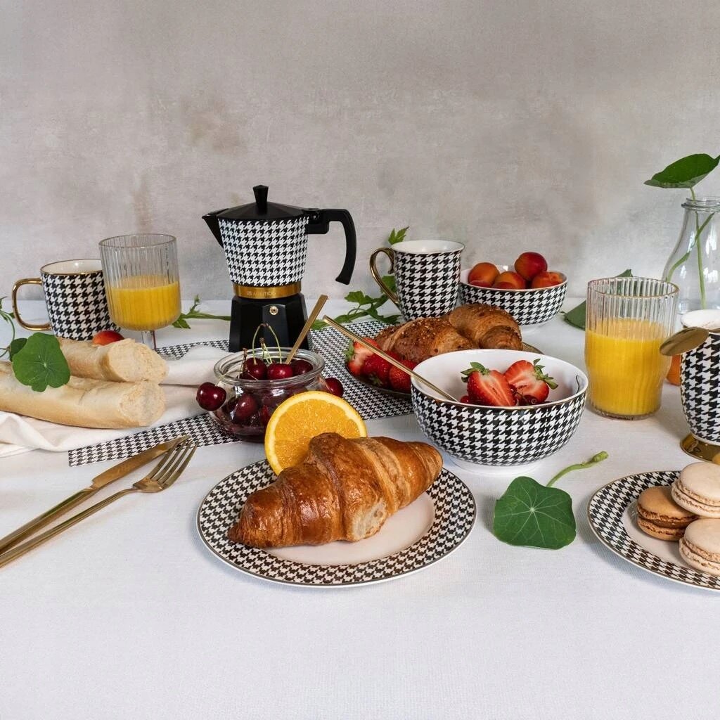 Na stole znajduje się zastawa w pepitkę, w tym kubki i miski, obok których leżą croissanty, owoce, bagietki i szklanki z sokiem pomarańczowym. W tle widoczny jest czajnik do kawy w podobnym wzorze.