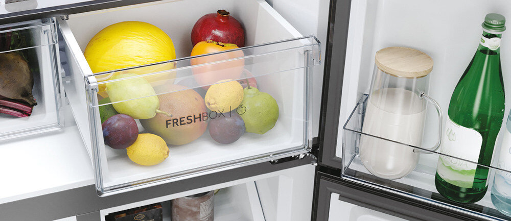 Wnętrze lodówki z otwartą szufladą oznaczoną napisem \'FRESH BOX\', w której znajdują się różne owoce. Obok w drzwiach lodówki widoczne są butelki i pojemnik z drewnianą pokrywką.