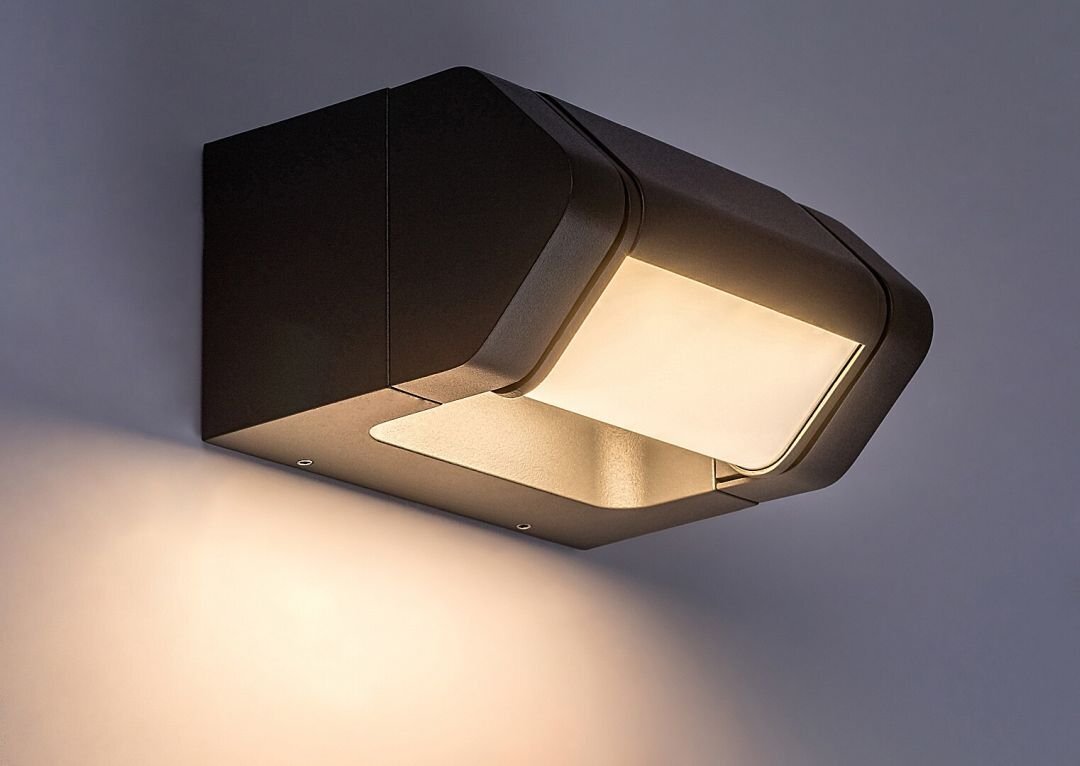 Na obrazie widoczna jest nowoczesna lampa ścienna w kolorze antracytowym, emitująca ciepłe światło. Lampa ma prostokątny kształt i jest zamontowana na ścianie.