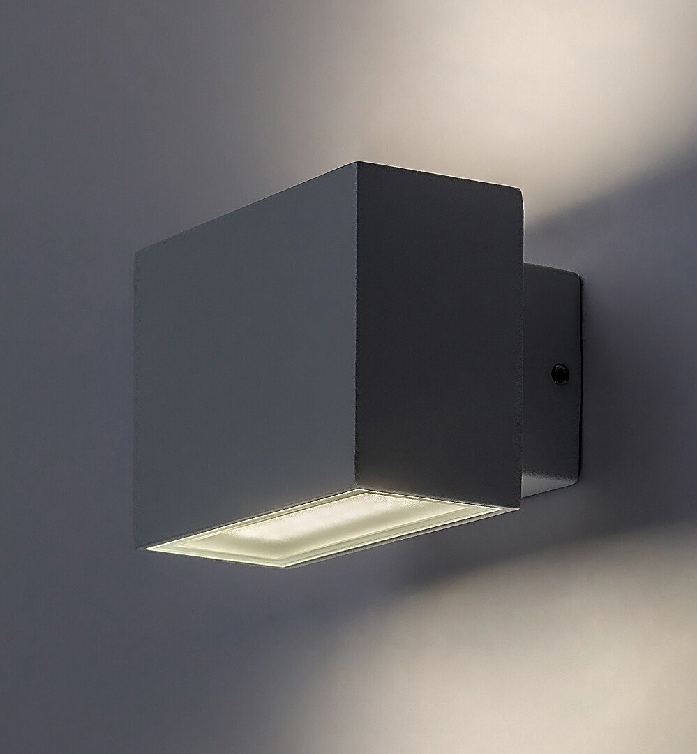 Na obrazie widoczna jest biała, aluminiowa lampa ścienna o kształcie prostopadłościanu, emitująca światło w górę i w dół. Lampa jest zamontowana na ścianie i oświetla powierzchnię wokół siebie.