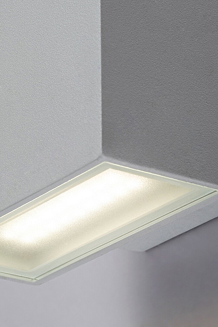 Biała lampa ścienna zewnętrzna LED zamontowana na ścianie, emitująca jasne światło. Lampa ma prostokątny kształt i jest wykonana z materiału przypominającego aluminium.