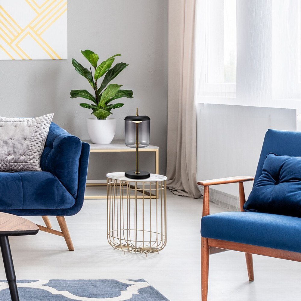 W nowoczesnym wnętrzu znajduje się lampa stołowa z czarną podstawą i szklanym kloszem na okrągłym stoliku z metalowym stelażem. Obok stoi niebieska sofa i fotel, a na stoliku w tle stoi roślina w białej doniczce.