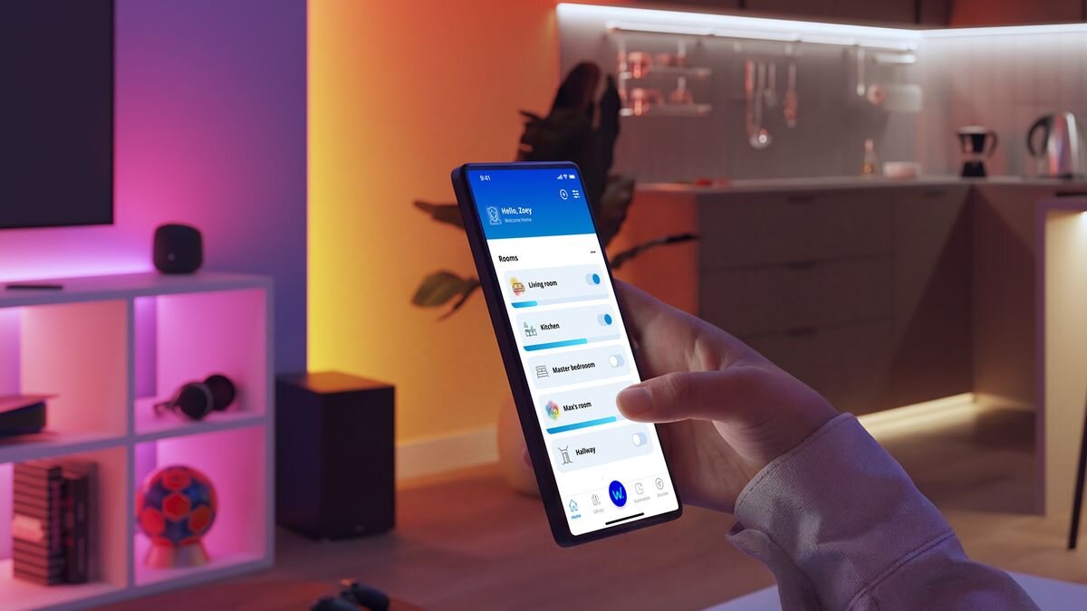 Osoba trzyma smartfon z aplikacją do zarządzania inteligentnym domem, na ekranie widoczne są różne pomieszczenia i urządzenia. W tle znajduje się nowoczesna kuchnia z kolorowym oświetleniem LED.