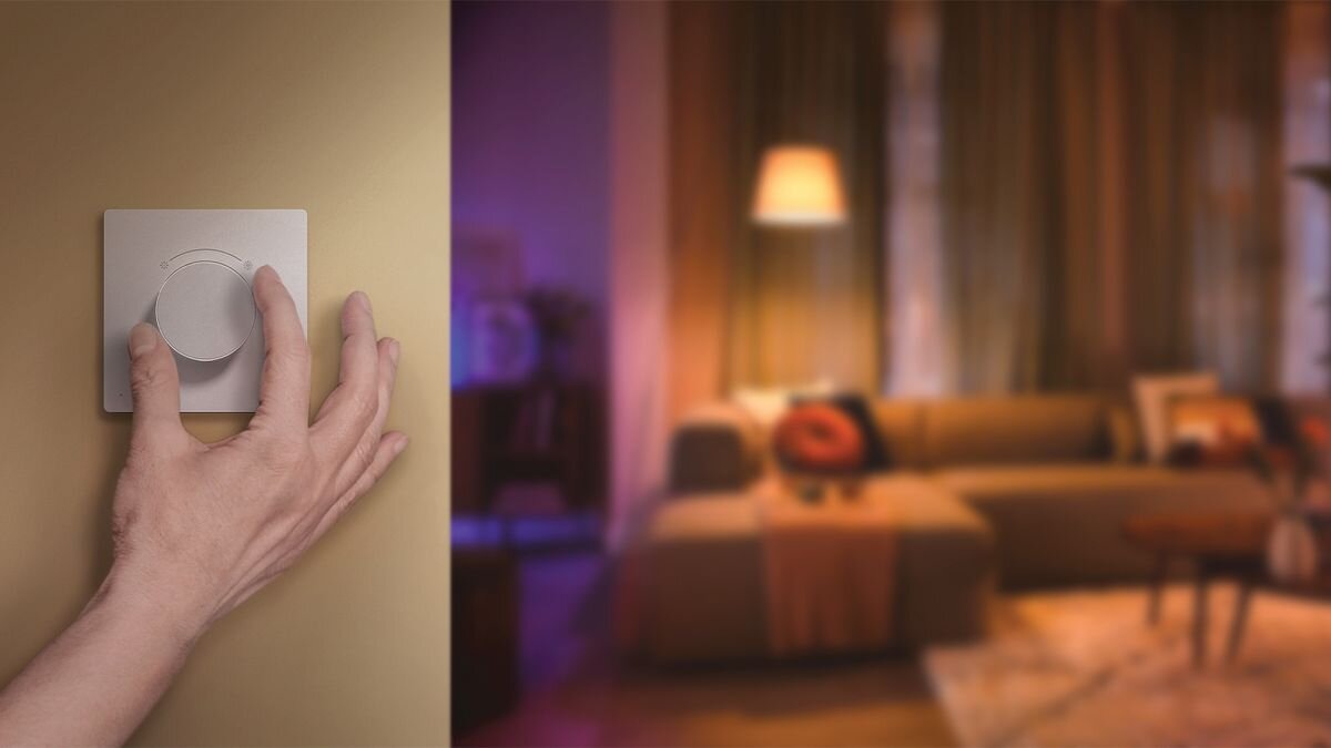 Ręka reguluje okrągły ściemniacz na ścianie, a w tle widać rozmyty salon z sofą i lampą.