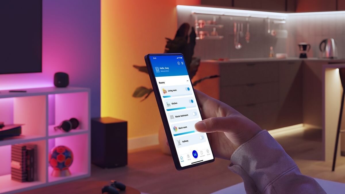 Osoba trzyma smartfon z aplikacją do sterowania oświetleniem, na ekranie widoczne są różne pomieszczenia i opcje. W tle znajduje się nowoczesna kuchnia z podświetleniem LED oraz regał z kolorowym oświetleniem.