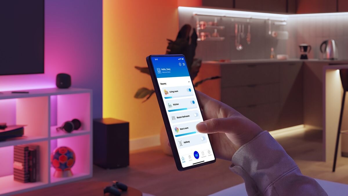 Osoba trzyma smartfon z aplikacją do sterowania oświetleniem, na ekranie widoczne są różne opcje pomieszczeń. W tle znajduje się nowoczesna kuchnia z podświetlanymi półkami i sprzętem AGD.