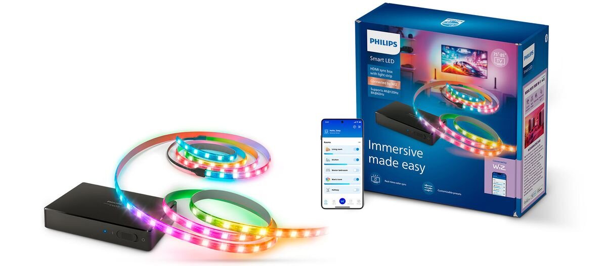 Na obrazie znajduje się zestaw z taśmą LED, czarnym urządzeniem i smartfonem z aplikacją, a obok widoczny jest niebieski karton z napisem \'PHILIPS Smart LED Immersive made easy\'.