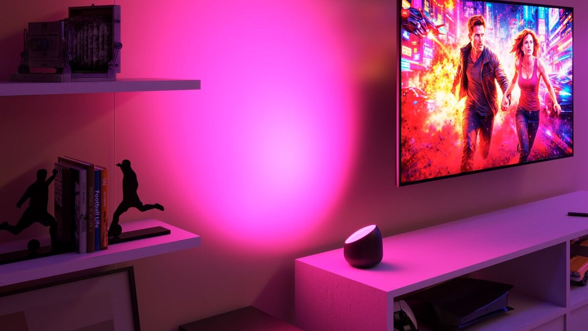 Pokój oświetlony różowym światłem z lampy stołowej, na półce znajdują się książki i figurki, a na ścianie wisi telewizor z dynamiczną sceną filmową.