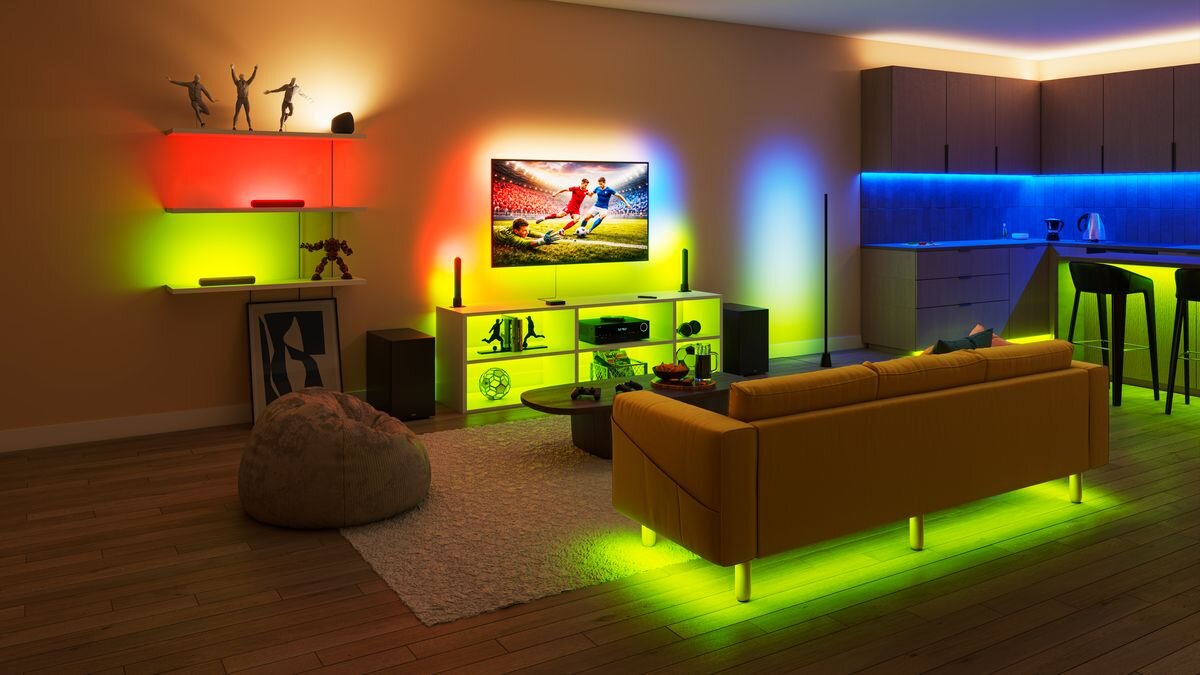 Pokój dzienny z nowoczesnym wystrojem, w którym znajduje się żółta sofa, telewizor z wyświetlanym meczem piłki nożnej oraz kolorowe oświetlenie LED na półkach i wokół mebli. W tle widoczna jest kuchnia z podświetlanymi szafkami.