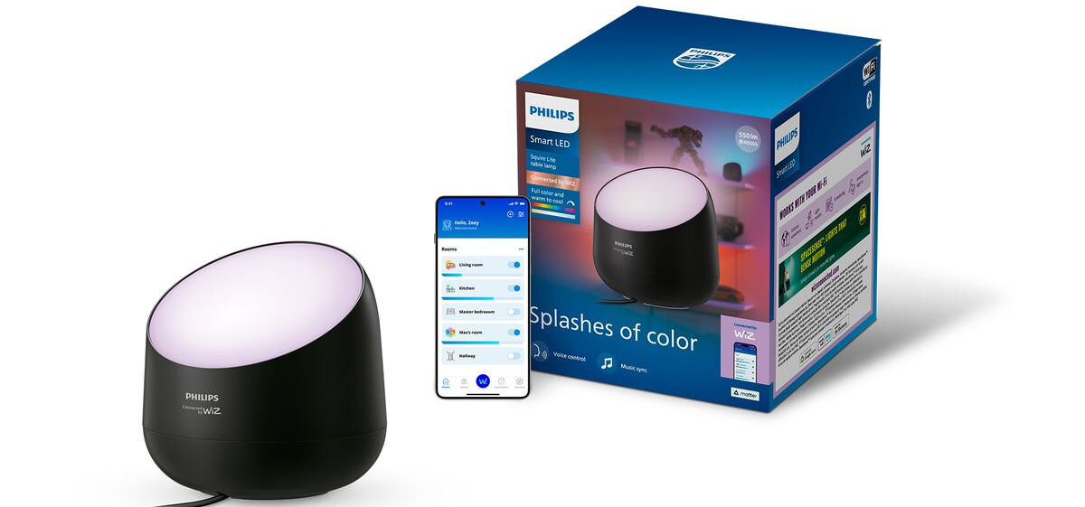 Na obrazie widoczna jest czarna lampa stołowa z białym, świecącym wierzchem oraz pudełko z napisem \'Splashes of color\'. Obok lampy znajduje się smartfon z aplikacją do sterowania oświetleniem.