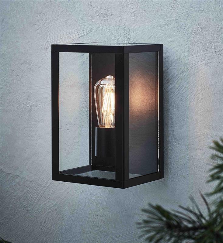 Czarna, prostokątna lampa ścienna z przezroczystymi panelami, wewnątrz której znajduje się żarówka o widocznym żarniku. Lampa zamontowana jest na jasnej, teksturowanej ścianie.