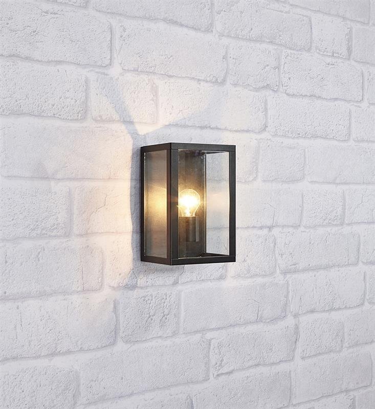 Na obrazie widoczna jest lampa ścienna w kształcie prostokątnej latarni z czarną ramą i przezroczystymi szybami, zamontowana na ścianie z białej cegły. Wewnątrz lampy znajduje się żarówka emitująca ciepłe światło.