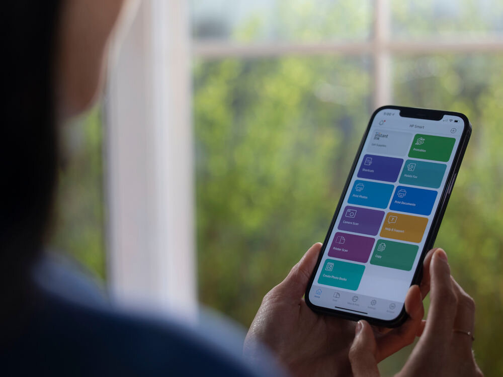 Osoba trzyma smartfon z aplikacją wyświetlającą kolorowe kafelki z różnymi funkcjami. W tle widoczne jest rozmyte okno z zielenią na zewnątrz.