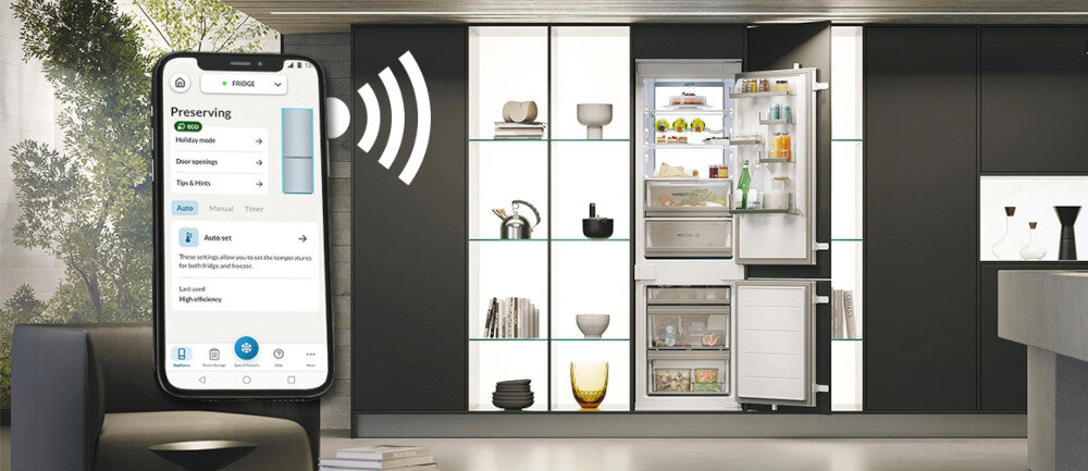 Wnętrze nowoczesnej kuchni z otwartą lodówką, w której widać różne produkty spożywcze. Obok lodówki znajduje się smartfon z aplikacją do zarządzania lodówką, na ekranie widoczny jest tekst: \'Preserving, Holiday mode, Door opening, Turn on FDD.\'