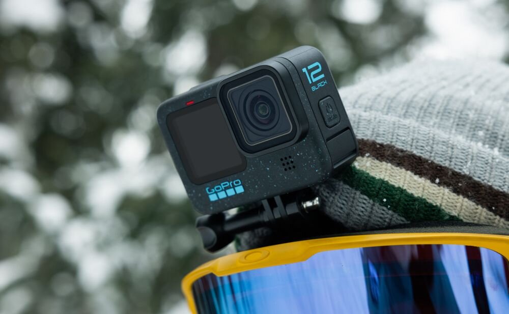 Kamera sportowa z napisem \'12 BLACK\' i logo \'GoPro\' zamontowana na kasku. W tle widoczne są rozmyte drzewa pokryte śniegiem.