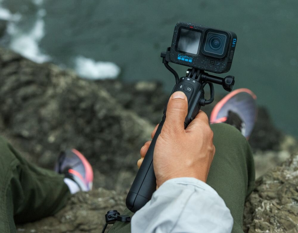 Osoba trzyma kamerę sportową zamontowaną na uchwycie, siedząc na skale nad wodą. Kamera jest czarna z widocznym logo \'GOPRO\'.