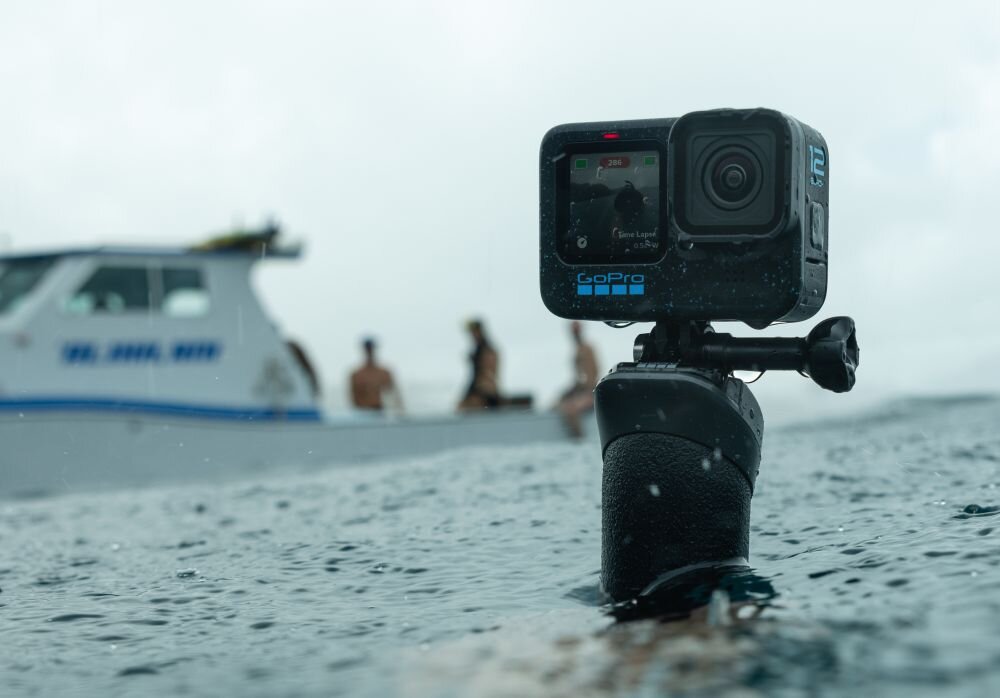 Kamera sportowa zamontowana na uchwycie wystającym z wody, w tle widoczna łódź i kilka osób. Na kamerze widoczne jest logo GoPro.
