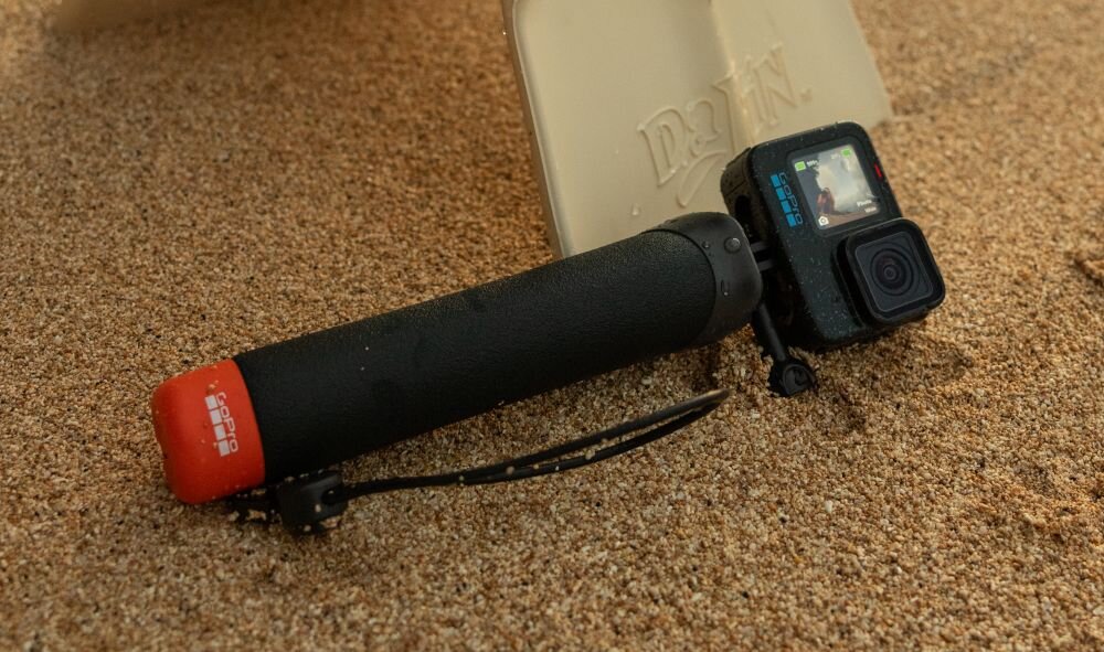 Kamera sportowa z czarnym uchwytem leży na piasku obok jasnego przedmiotu z napisem \'DRIFT\'. Na uchwycie widoczny jest czerwony element z białym logo \'GOPRO\'.