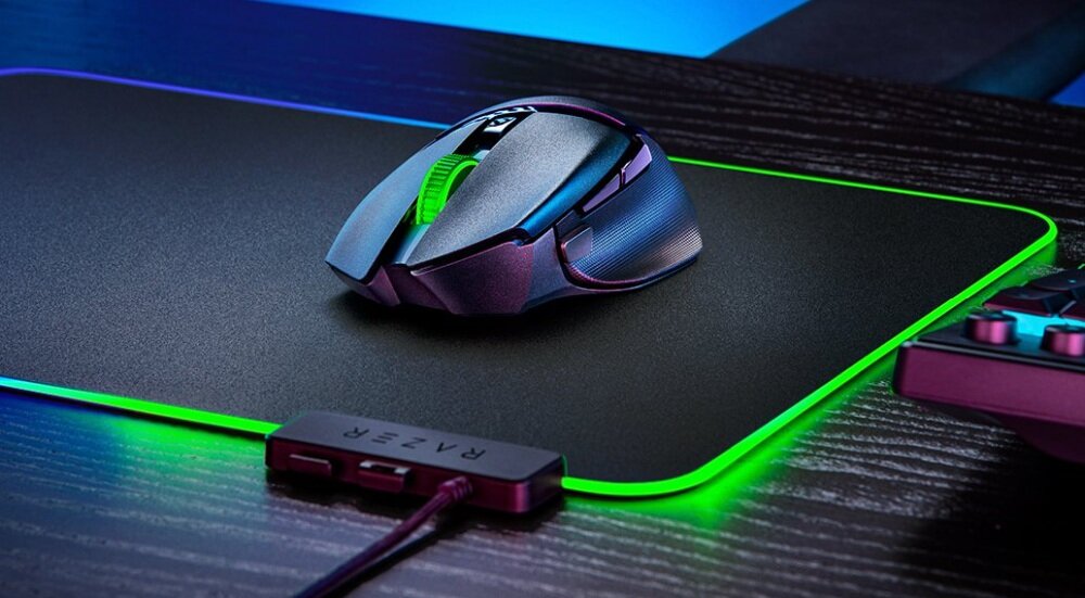Na czarnej podkładce z zielonym podświetleniem leży mysz komputerowa o nowoczesnym designie. Obok widoczny jest element z logo \'RAZER\'.