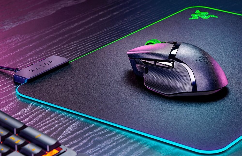 Na obrazie widoczna jest czarna mysz komputerowa z podświetleniem RGB, umieszczona na podkładce z logo RAZER. Obok znajduje się urządzenie z napisem \'RAZER\'.