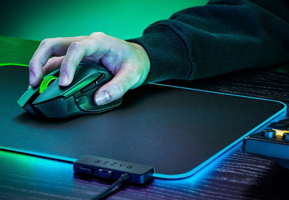 Osoba trzyma czarną mysz komputerową z zielonym elementem na podkładce z podświetleniem. Na podkładce widoczne jest logo \'RAZER\'.