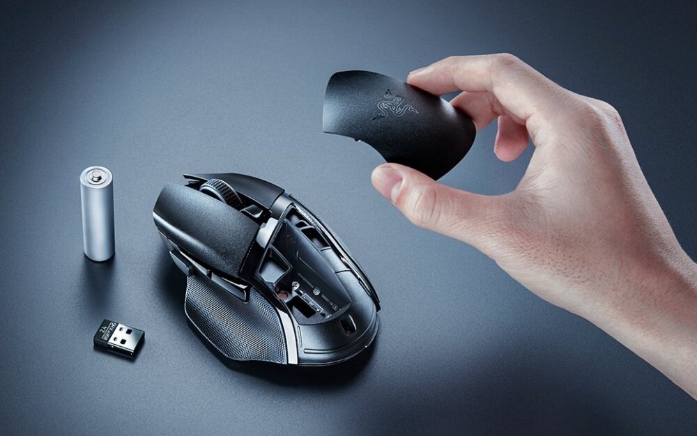 Na ciemnym tle widać mysz komputerową z otwartą obudową, obok której leży bateria i mały odbiornik USB. Ręka trzyma zdejmowaną część obudowy z logo w kształcie trzech węży.