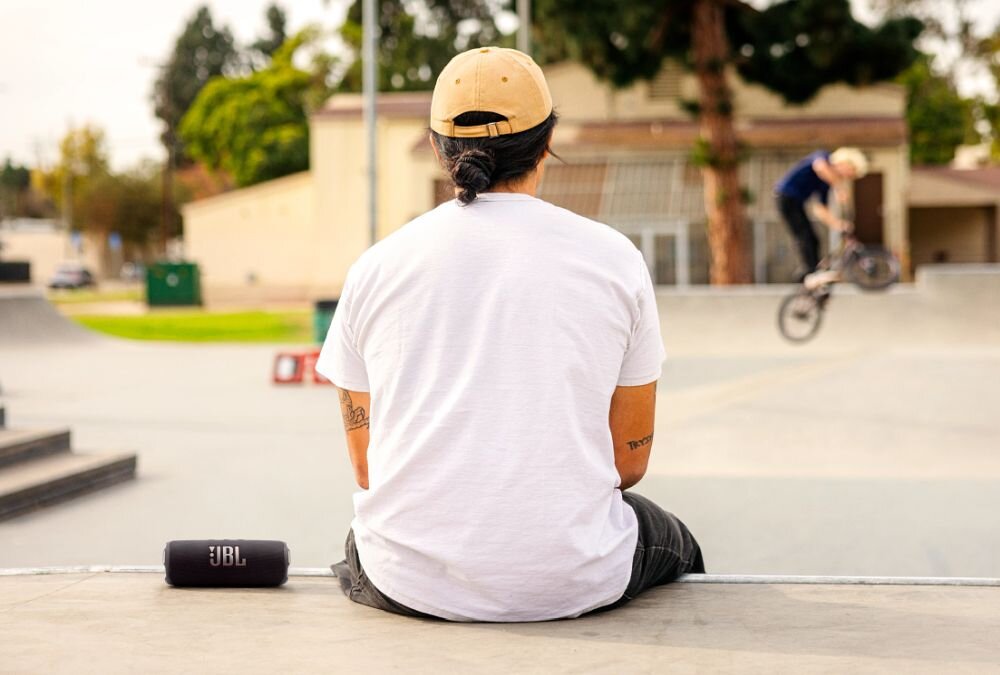Osoba w białej koszulce i beżowej czapce siedzi na rampie w skateparku, obok znajduje się fioletowy głośnik z logo JBL. W tle widać osobę na rowerze BMX wykonującą trik.